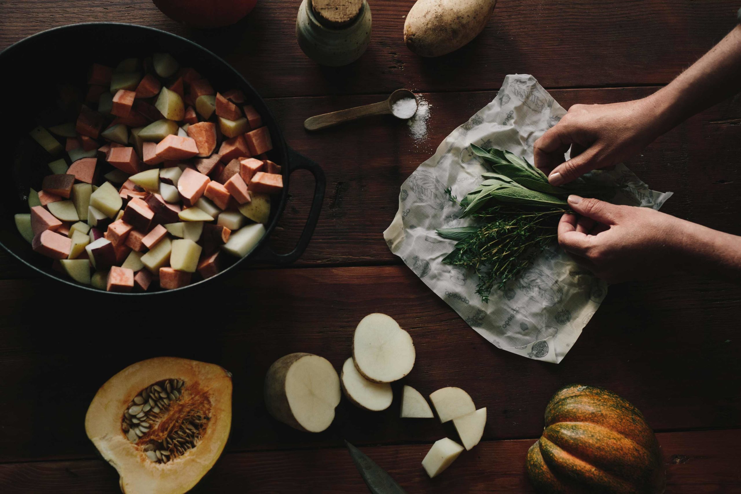 Overhead shot of a wooden table with a large cast iron pot with cut up root vegetables, half a squash, and sliced potatoes. There is an open package of Abeego wax food wrap containing herbs