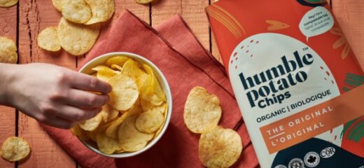A picture of a hand picking chips from a bowl full of humble potato chips and a Humble potato chips packet & some chips are spreaded on the surface.