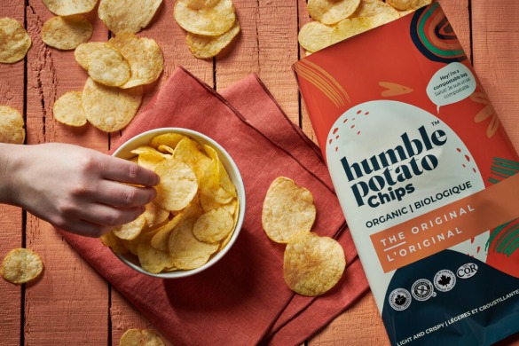 A picture of a hand picking chips from a bowl full of humble potato chips and a Humble potato chips packet & some chips are spreaded on the surface.