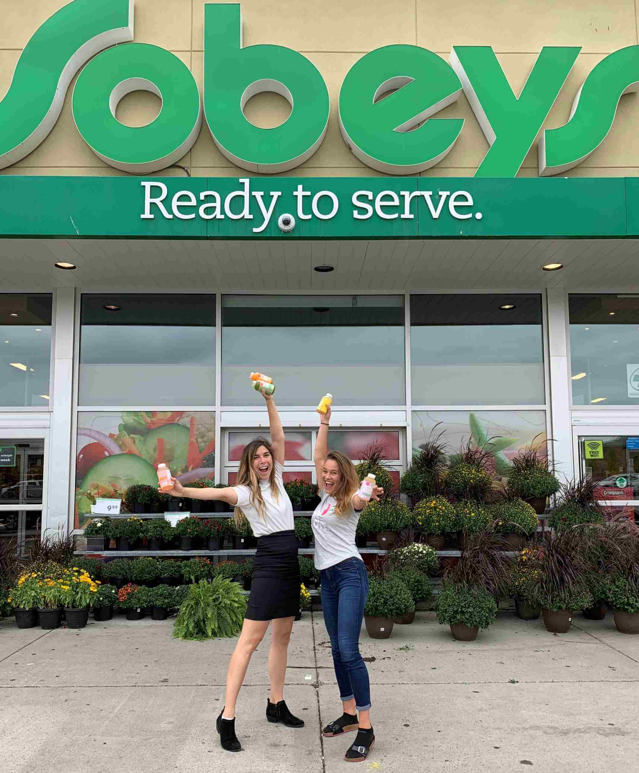The image depicts two women taking a picture in front of the 'Sobeys Ready to Serve' store while holding bottles of drinks in their hands.